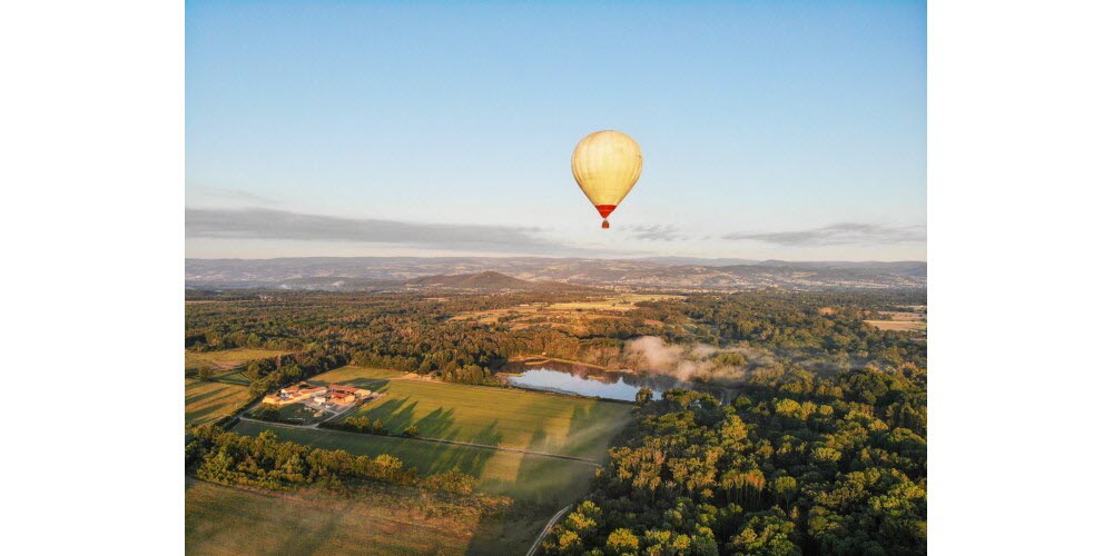 Vue aérienne d'une montgolfière survolant la plaine du Forez au lever du soleil : champs, étang, ferme et montagnes à l'horizon